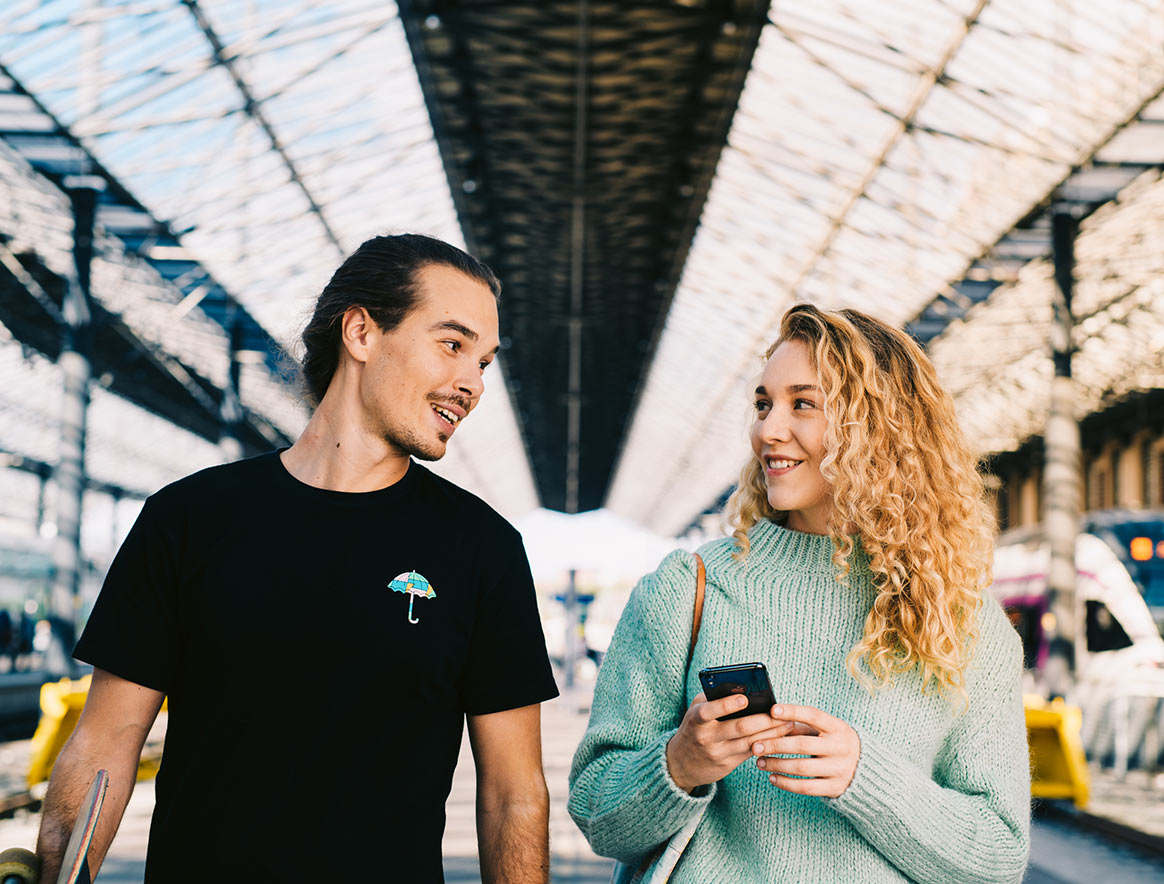 Man and a woman on a railway station walking and talking. Woman is holding a phone.